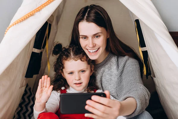 Positive young mum using social media app on smartphone recording video blog having fun with cute small kid girl embracing bonding together looking at mobile phone, sitting in cozy tent at home