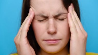 Stresli, genç, esmer bir kadının yatışmış portresi baş ağrısını dindiriyor, stres atıyor, yorgun, sinirli, masaj yapan, mavi stüdyo arka planında izole edilmiş bir kız.