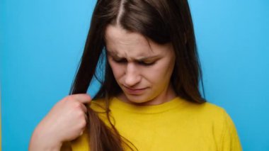 Depresyondaki genç bir kadının hasarlı saçlara dokunuşunun yakın portresi, saç dökülmesinden endişe etmek, mavi stüdyo arka planında izole edilmiş, hormonal dengesizlikten muzdarip, kameraya mutsuz bakan.