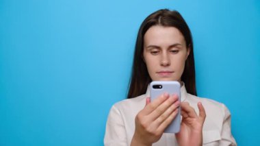 Güzel sakin esmer kızın portresi cep telefonuyla duruyor, beyaz gömlek giyiyor, boş alanda işaret parmağı ile işaret ediyor, mavi stüdyo arka planında izole edilmiş. İnsanlar, din, teknoloji