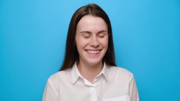 幸せなブルネットの女性の肖像画は喜んで笑い 陽気な肯定的な若い女性は 青いスタジオの背景に隔離された白いシャツに身を包んだ誠実な感情を表現します 人々とライフスタイルのコンセプト — ストック動画