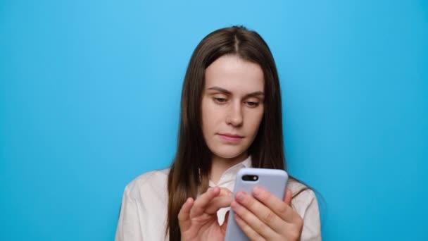 兴奋的黑发女人在网上大获全胜 兴奋的年轻可爱女人在智能手机上读到意想不到的好消息 身穿白衬衫 与蓝色工作室隔离 — 图库视频影像