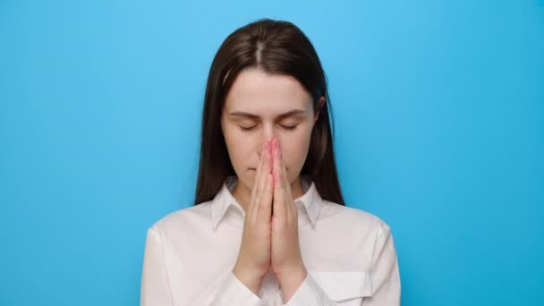 希望の迷信的な女性の肖像画の祈りの中で手を維持し 若い宗教的な女の子の信者の祈り 希望を求める最高の 白いシャツを着て 青いスタジオの背景に隔離された 信仰の概念 — ストック動画