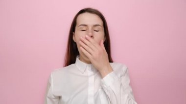 Uykulu, genç bir kadın esniyor, uykulu, ilgisiz, uykusuz, tembel, uykusuzluktan sıkılmış, beyaz gömlek giymiş, pembe arka planda soyutlanmış. Yorgunluk ve uyanış kavramı