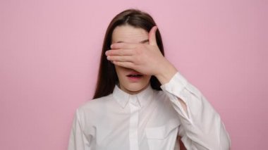 Genç kız yüzünü eliyle kapatıyor, ağzını açıyor, birinden saklanmaya çalışıyor, kadın şok oluyor, gözleri kapanıyor, sürpriz için bekliyor, beyaz gömlek giyiyor, pembe stüdyo arka planında soyutlanıyor.