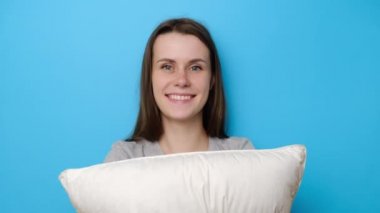 Neşeli esmer kadın yastığı kucaklıyor, kameraya sevinçle bakıyor, geniş gülümsüyor, mutlu genç kız tembellikten ve gevşemeden hoşlanıyor, mavi stüdyo arka planında soyutlanmış. Uyanış konsepti.