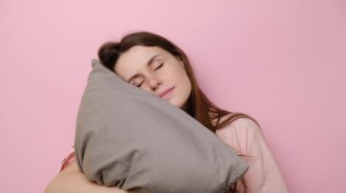 Neşeli genç bir kadın yastıkla uyanır, yanaklarına dokunur, kameraya bakarak mutlu bir şekilde yaşar, pijama giyer, sağlıklı bir uykudan sonra iyi bir ruh hali içinde olur, pembe stüdyo duvarında izole olur. Uyku konsepti