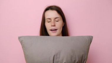 Uykulu, neşeli, genç bir kadının portresi gri yastığı kucaklıyor, pijama giymiş bir şekilde uyumak istiyor, terfiniz için boş bir arkaplan ile mavi stüdyoda poz veriyor. Uyku konsepti