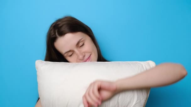 快乐的女人带着枕头醒来 触摸着脸颊 快乐地看着镜头 穿着T恤衫 在健康的睡眠后心情很好 与蓝色背景隔离 睡觉的概念 — 图库视频影像