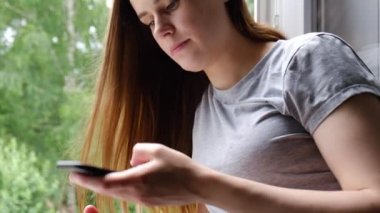 Genç esmer kadın pencere pervazında oturmuş telefonla konuşuyor. Sanal dijital ses asistanını aktive ediyor. Kız arkadaşlarıyla internetten ya da hoparlörden konuşuyor.