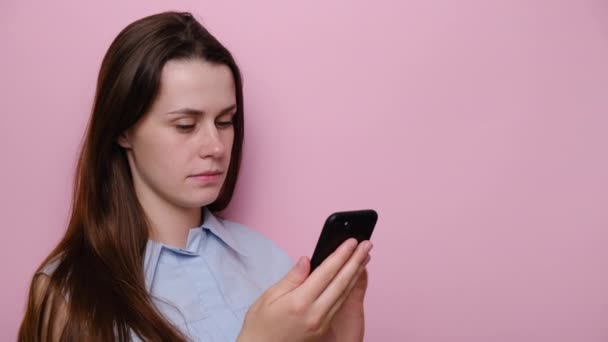 在智能手机上看到坏消息 穿着蓝色衬衫 在粉红的工作室背景下被隔离的悲伤小女孩的画像 惊慌失措的黑发女人心情不好 忧心忡忡的女人检查电话内容 — 图库视频影像