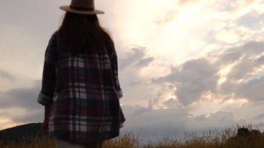 Şapkalı genç bir kadının güzel bir günbatımında arka plandaki dağda yürüdüğü arka plan görüntüsü ellerini havaya, gençliğe ve mutluluğa kaldırıyor. Gerçek vahşi yaşam tarzını keşfet.