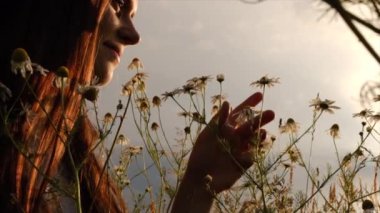 Şapkalı beyaz kadın gün batımında zevkten gözleri kapalı papatyaları kokluyor. Çiçeklerin peri çayırında gülümseyen kızın mutlu romantik portresini kapat. Yavaş çekim