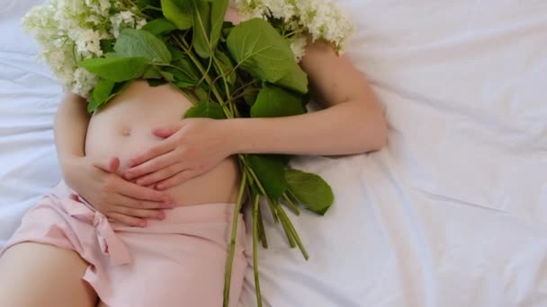 美しい妊婦のトップビューは優しく新鮮な花束の花と自宅で居心地の良い白いベッドの上に横たわるおなかの手をストロークします 将来の母親の期待 コンセプト 母子家庭 — ストック動画