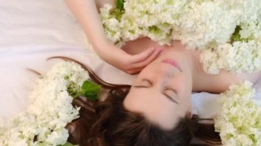 Romantik, genç, çekici, taze çiçekli, rahat beyaz bir yatakta uzanmış, nazikçe yüzünü okşayan, mutlu bir şekilde kameraya bakan bir kadının üst görüntüsü. Gülümseyen, yaratıcı, şehvetli esmer kız portresi