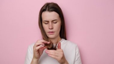 Kırık uçlara bakan depresif genç bir kadın, pembe stüdyo arka planında izole edilmiş. Kadın hormon problemleri ya da vitamin eksikliği. Mutsuz kız hasara uğramış kuru saç dökülmesi konseptine üzülüyor.
