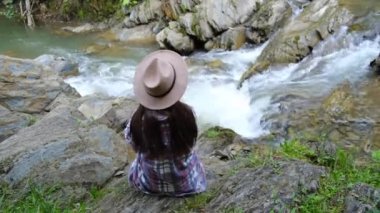 Genç hippi kadın ya da şapkalı bir gencin kayanın üzerinde oturup güçlü dağ suyu akıntısına ya da yazın akan nehre bakması. Kavramsal dünya gezgini, seyahat tutkusu