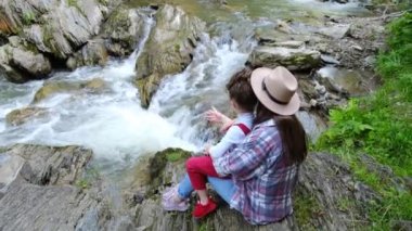 Şapkalı sakin bir anne ve küçük tatlı kızının yaz boyunca küçük bir şelale üzerinde manzaranın tadını çıkarması. Aile macerası konsepti, açık havada doğal harikaları keşfetmek.