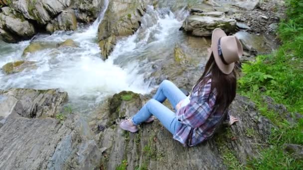 年轻的孕妇头戴礼帽坐在悬崖边上 望着汹涌澎湃的山水流淌 美丽的自然景观 安静的绿色旅游胜地 概念世界旅行者或博主 游手好闲 — 图库视频影像
