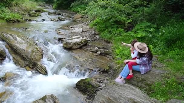 野生の川の近くの崖の端に座っている帽子の幸せなお母さんとかわいい未就学児の娘は 美しい夏の景色を楽しんでいます 夢と感動的な家族の中を歩く アウトドアアクティビティ旅行のコンセプト — ストック動画