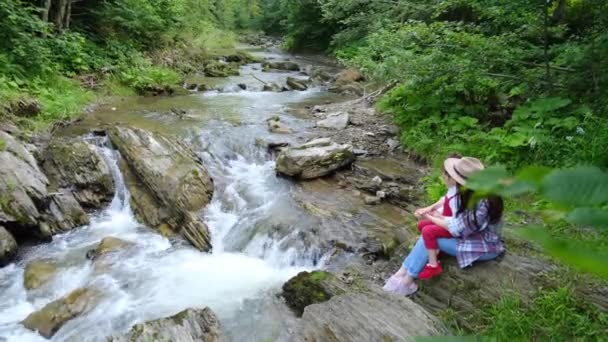 戴着帽子的快乐的年轻女子和可爱的学龄前女儿坐在悬崖边欣赏喀尔巴阡山脉风景中美丽的小瀑布 概念冒险家庭探索旅游博客 — 图库视频影像