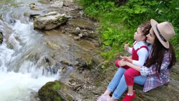 石の急流と水しぶきで川の近くに彼女の小さな未就学児の娘と一緒に座ってかわいい若い母親 幸せな家族は都市の外の週末に一緒に時間を過ごす アクティブレクリエーションコンセプト — ストック動画
