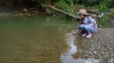 Şapkalı genç bir anne ve küçük tatlı bir kız ormanın şeffaf nehrinde nazikçe suyun yüzeyine dokunuyor. Doğayla barışçıl ve düşünceli bir bağ. Ailenin açık hava eğlencesi hariç.
