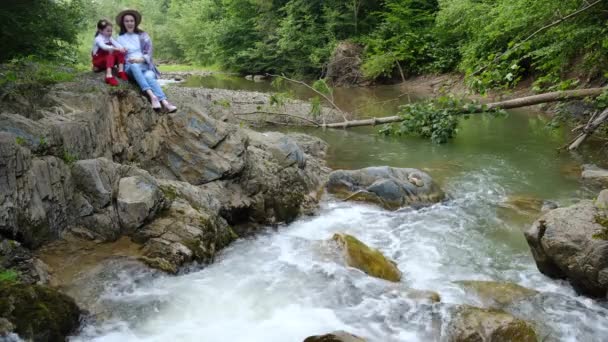 快乐的年轻妈妈戴着帽子 可爱的小女儿坐在湍急的河边悬崖边 欣赏美丽的夏日美景 梦想和鼓舞人心的家庭度假 活动旅行概念 — 图库视频影像