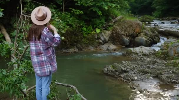 边景迷人的戴帽子的女人 站在山河边 举手投足 青春快乐 可爱的女孩欣赏美丽的森林风景 探索真正的野外生活方式 — 图库视频影像
