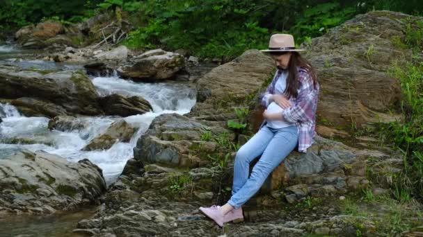 年轻美丽的孕妇头戴礼帽 呼吸新鲜空气 真实而自然的美丽形象 凝视着碧绿森林的迷人自然景观 以及闪烁着石花的河水 — 图库视频影像