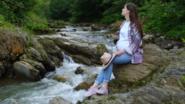美丽的孕妇坐在靠近野生河流的悬崖边 拍打着肚子 享受着令人难以置信的宁静的森林 呼吸着新鲜空气 在风中吹拂着头发 自然奇观 安静的旅游胜地 — 图库视频影像