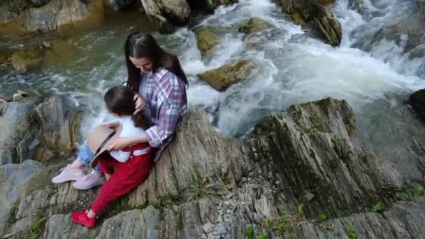 陽気妊娠中の母親と少女山の川の近くのエッジの石の上に座って お母さんと小さな娘が一緒にリラックスし 柔らかい瞬間をお楽しみください 心からの感情や積極的なレクリエーションを屋外で — ストック動画