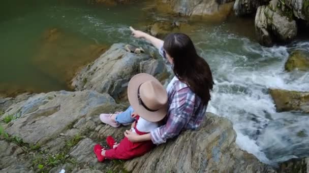 穿着衬衫的年轻的黑发母亲和戴着帽子的可爱的学龄前女儿坐在溪流或河流边的悬崖边 俯瞰着森林中的山下小山 探索旅行的冒险家庭概念 — 图库视频影像
