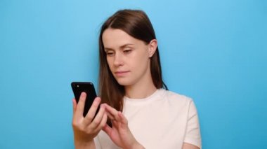 Akıllı telefonlu esmer kadının portresi yakın plan. İyi haber içeren mesajlar alıyor. Mavi stüdyo arka planında izole edilmiş evet hareketi yapıyor. Samimi duygular gösteren zafer anı