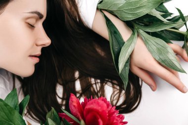 Beyaz stüdyo arka planında kırmızı şakayıklar ve yeşil yapraklar olan rüya gibi esmer bir kadının güzel portresi. Güzel, güzel, temiz ciltli kızın yüzünü kapat.