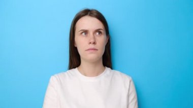 Düşünceli genç bir kadın, zor bir seçim üzerine düşünürken, beyaz tişört giymiş, üzerinde kafa yoran, bilgisiz görünen, mavi stüdyo arka planında izole edilmiş bir plan hayal ederken,