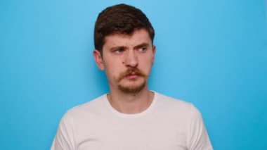 Mutsuz milenyum adamı parmaklarıyla burnunu sıkıyor, berbat bir şey kokuyor, iğrenç bir ifadeyle kameraya bakıyor, tişört giyiyor, kötü koku hakkında kötü hissediyor, mavi stüdyo arka planında izole edilmiş.