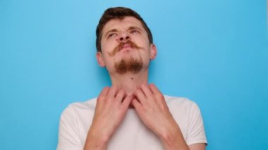 Alerji belirtileri gösteren, boynu kaşınan, beyaz tişörtlü, mavi stüdyo arka planında fotokopi alanı olan sağlıksız üzgün bin yıllık bir adamın portresi. Sağlık sorunu ve deri hastalıkları kavramı