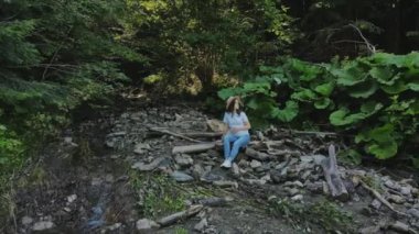 Şapkalı hamile bir kadının hava aracı görüntüsü. Küçük nehir suyunun yanındaki yeşil ormanda vakit geçirir. Oturur, rahatlar, doğanın ve temiz havanın tadını çıkarır. Hafta sonu kaçamağı ya da kamp gezisi