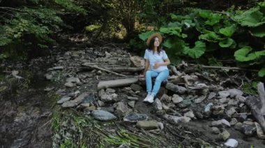 Şapkalı genç ve güzel bir kadının hava aracı görüntüsü küçük nehir suyunun yakınındaki yeşil karışık ormanda vakit geçirir, oturur ve rahatlar, doğanın ve temiz havanın tadını çıkarır. Macera kavramını ve seyahat günlüğünü keşfetmeyi