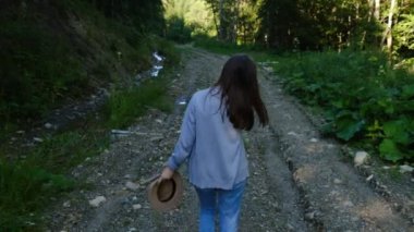Genç esmer bir kadının arkadan görünüşü, kot pantolon ve şapka giyiyor, yeşil ormanın yakınındaki dağ yolunda gelişigüzel yürüyor, doğanın tadını çıkarıyor. Seyahat tutkusu blogcularına ilham vermek. Seçici odaklanma. Yavaş çekim