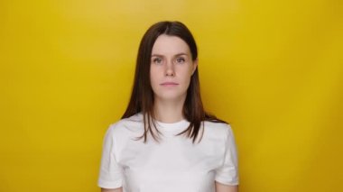 Hayal kırıklığına uğramış genç bir kadın, yüzünü eliyle kapatıyor, esmer kız kendini umutsuz hissediyor, stres ve gerginlikten muzdarip, sarı stüdyo arka planında izole edilmiş beyaz tişört giyiyor.