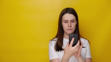 Beyaz gömlekli, sarı stüdyo arka planında izole edilmiş akıllı telefondan kötü haberler alan bin yıllık bir kadın. Şok olmuş genç bayan kendini kötü hissediyor, endişeli kız telefon içeriğini kontrol ediyor.