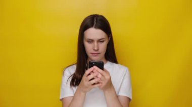 Mutlu bir kadın akıllı telefonu internet uygulamalarıyla tutuyor, gülümseyen tatlı bir kız telefonda mesajlaşıyor cep telefonuna bakıyor, beyaz tişört giyiyor, sarı stüdyo arka planında izole edilmiş.