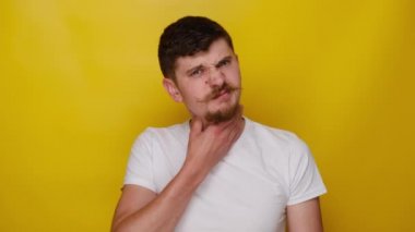 Yorgun adam ellerini boynundan ayırmıyor, boğularak ölüyor, boğazı ağrıyor, rahatsız oluyor, sağlıksız görünüyor, kameraya mutsuz bakıyor, yutması zor, sarı stüdyo arka planında izole edilmiş.