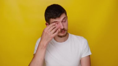Burun köprüsüne masaj yapan üzgün, göz yorgunluğu çeken, baş ağrısı ya da migrenden muzdarip, beyaz tişörtlü, sarı arka planda izole edilmiş genç bir adam. Gözler yorgunluğu konsepti