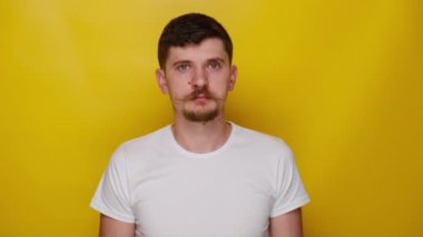 Komik, çekici bir genç adamın portresi kaşlarını çatıyor, kameraya bakıyor, beyaz t-shirt giymiş, sarı stüdyo arka planında izole edilmiş,