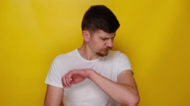 Dirsek ağrısı çeken mutsuz genç bir adam, kol kasları ağrılı, beyaz tişört giymiş, sarı stüdyo arka planının üzerinde duruyor. Ağrı kesici ve kronik eklem romatizması konsepti