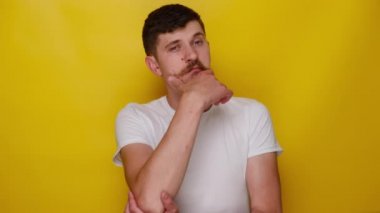 Düşünceli, kafası karışmış, çenesine dokunan, çözümü düşünürken başını kaşıyan, zor seçimler hakkında şüpheleri olan, sarı arka planda izole edilmiş tişört giyen genç bir adamın portresi.