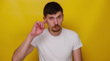 Şok olmuş genç bir adam sarı stüdyo arka planında aptal jestler yapıyor ve kamerayı işaret ediyor, çılgın bir kaçığı suçluyor, pervasız plan yapıyor, çılgın fikirleri suçluyor, beyaz tişört giyiyor..